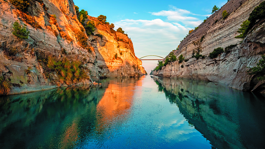 Narrow waterway between steep rocky cliffs with calm blue water and a distant bridge under a bright sky.
