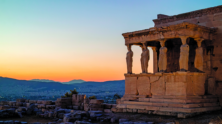 Ancient stone temple with sculpted columns shaped like standing figures, overlooking ruins and distant hills at sunset.