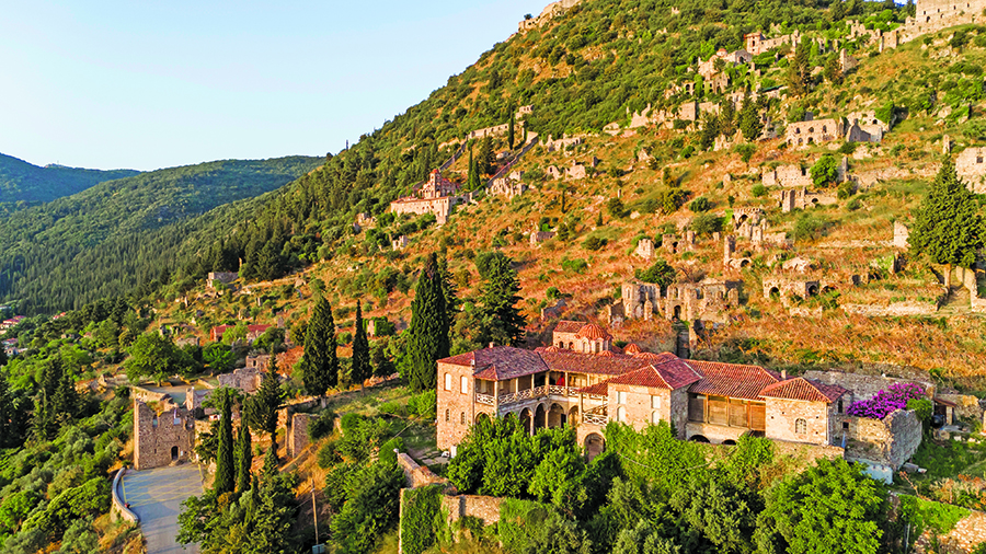 Stone buildings with red roofs nestled on a green hillside surrounded by trees and terraced slopes.