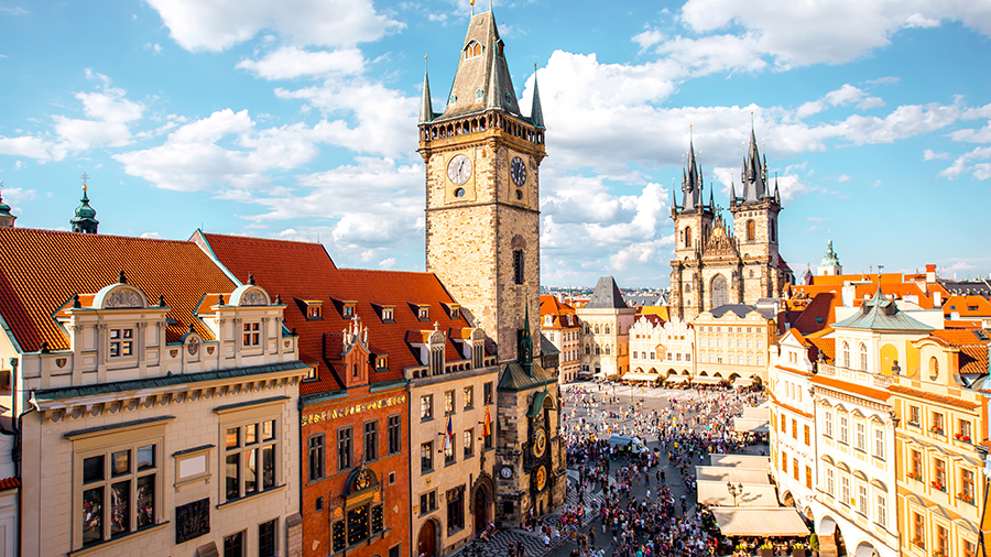 A busy historic city square filled with people, surrounded by colorful buildings and tall towers under a bright sky.
