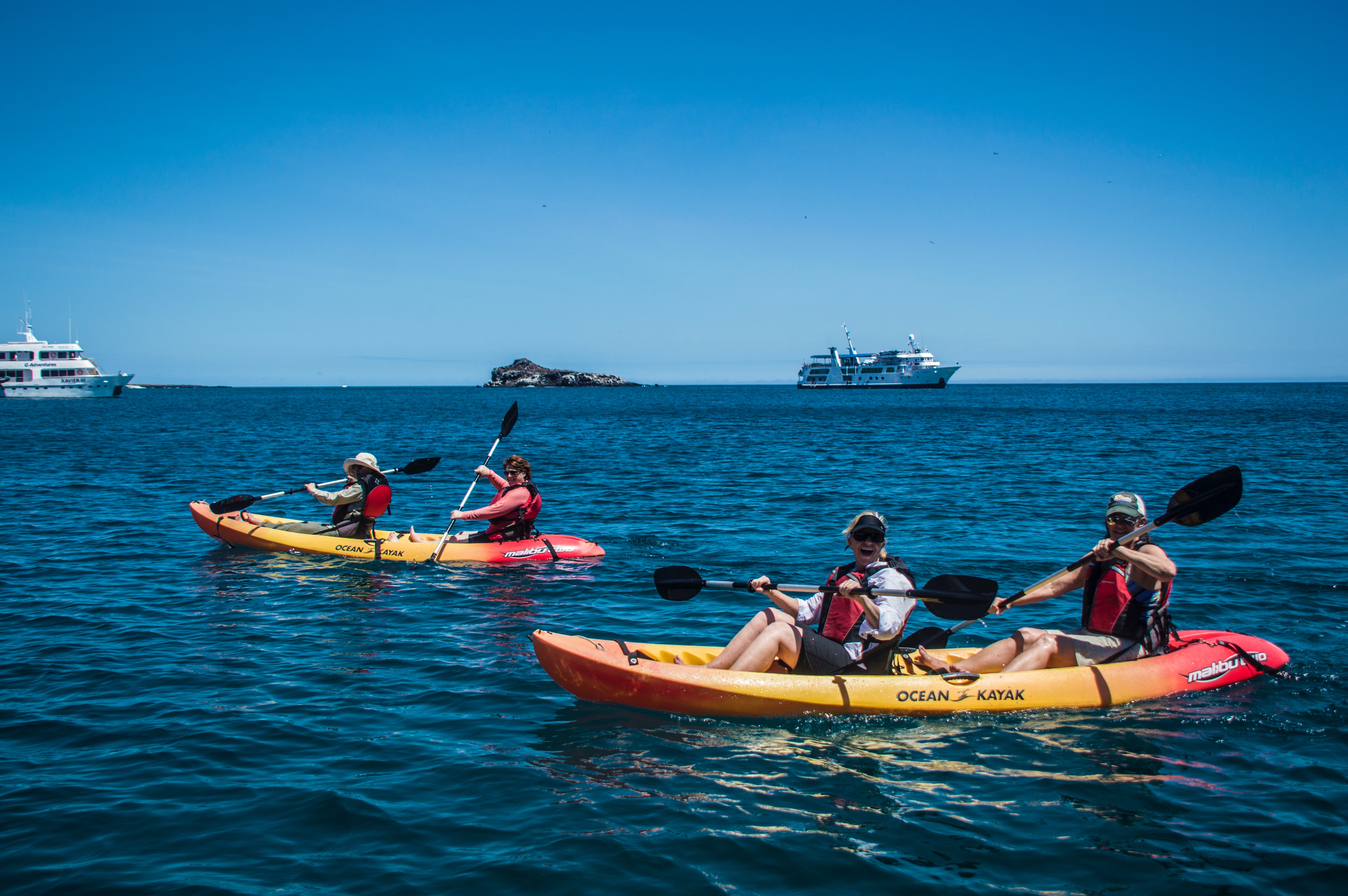 Two kayaks with people out in the water with boats in the background.