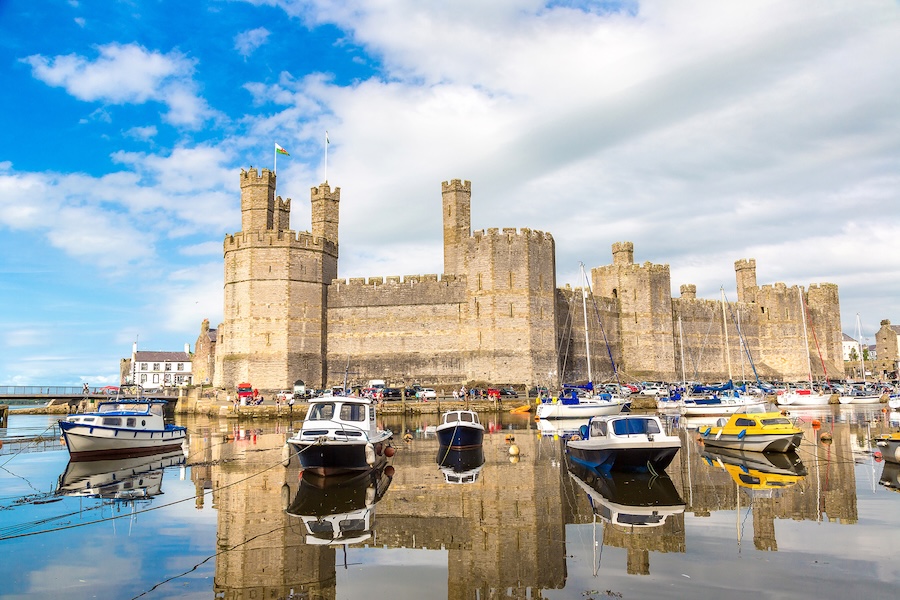 A historic stone castle with multiple turrets sits by a calm harbor under a partly cloudy sky. Colorful boats are moored, reflecting in the water.