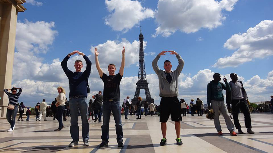Group of people with arms raised spelling O-H-I-O in an outdoor plaza, with the Eiffel Tower and a partly cloudy sky in the background.