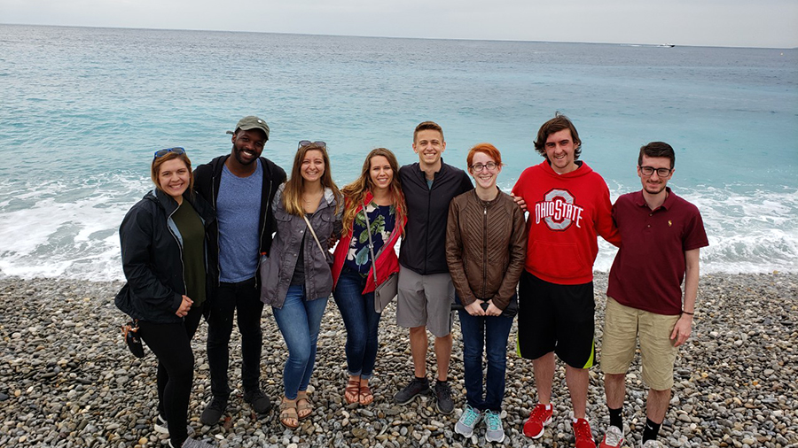 Group of people standing side by side on a rocky beach in front of the ocean, with waves behind them and an overcast sky.