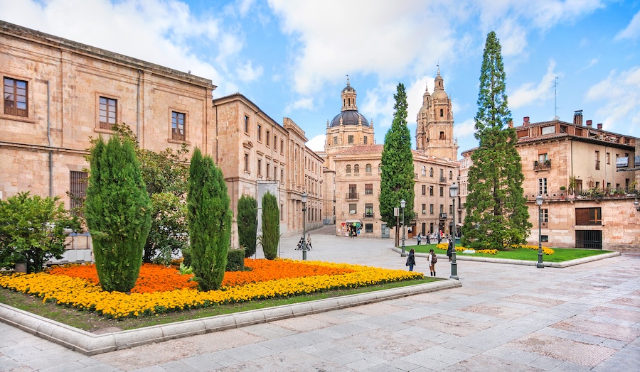 A vibrant square with colorful flower beds, surrounded by historic buildings and tall trees, under a partly cloudy sky.