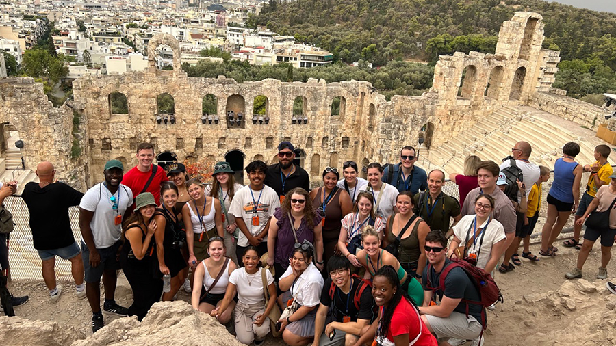 Large group of people posing together on stone steps inside ancient ruins, with arched walls and rocky hills in the background.