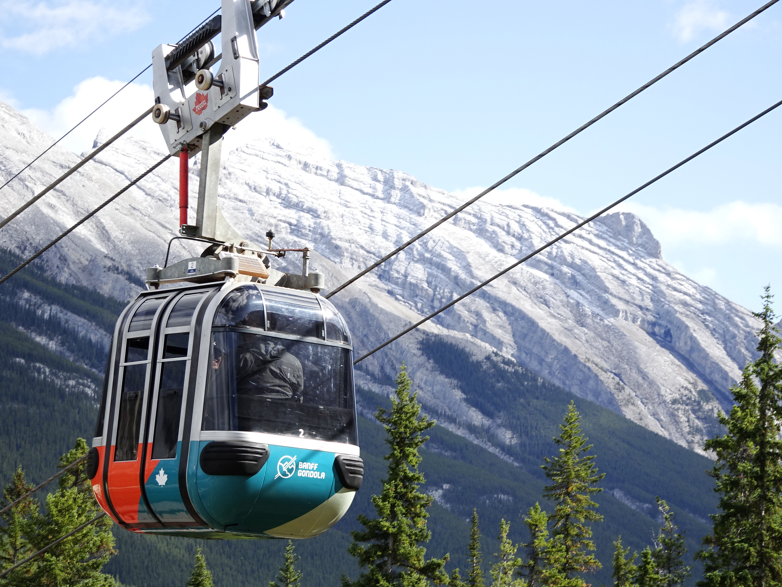 A Banff Gondola cabin rising above evergreen forests with the Canadian Rockies towering in the background.