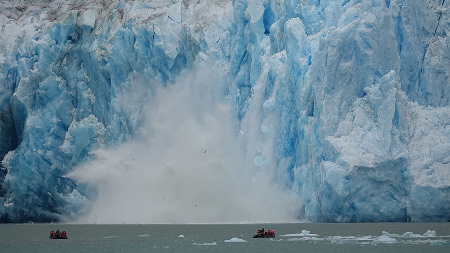 A massive blue glacier towers over the water as ice breaks off and crashes into the sea, creating a large splash near small boats below.