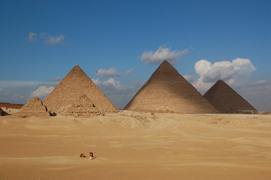 Three large pyramids rise from a sandy desert under a blue sky with scattered clouds.