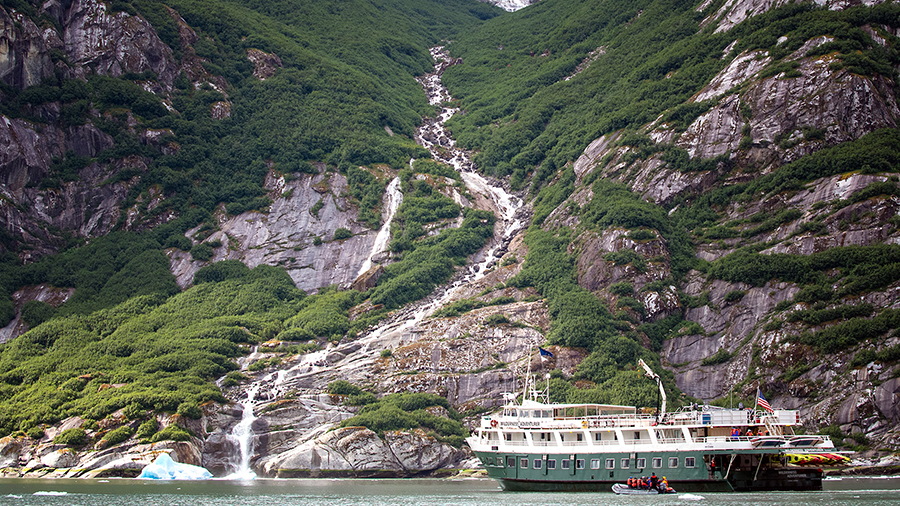 A white tour boat floats on calm water near a steep, green mountainside with a tall waterfall cascading down rocky cliffs into the sea.