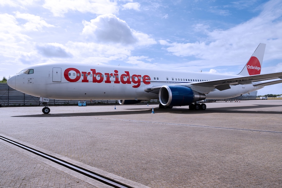 A white passenger airplane with the red “Orbridge” logo is parked on an airport runway under a partly cloudy blue sky.