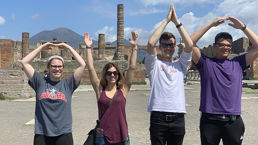 Four people standing in ancient ruins with stone columns behind them, each raising their arms spelling O-H-I-O under a blue sky.