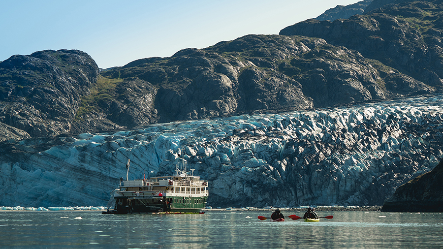 A tour boat floats on calm water in front of a large blue glacier backed by rocky mountains.