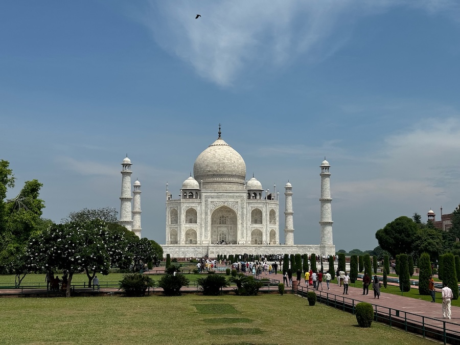 The Taj Mahal stands centered in a landscaped garden, with its white marble dome and four minarets under a blue sky, as visitors walk along the main pathway in front of the monument.