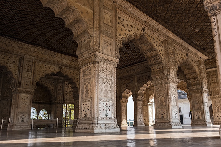 An ornate stone hall with carved arches and pillars, featuring detailed floral and geometric patterns on the columns, walls and ceiling, with sunlight reflecting on the polished floor.