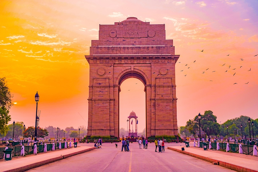 A large stone arch monument stands at the center of a wide road, with people walking beneath it, trees and streetlights on both sides and a warm orange and pink sky in the background.