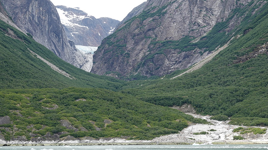 A wide green valley stretches between steep rocky mountains, with a winding river in the foreground and a glacier visible at the far end of the valley.