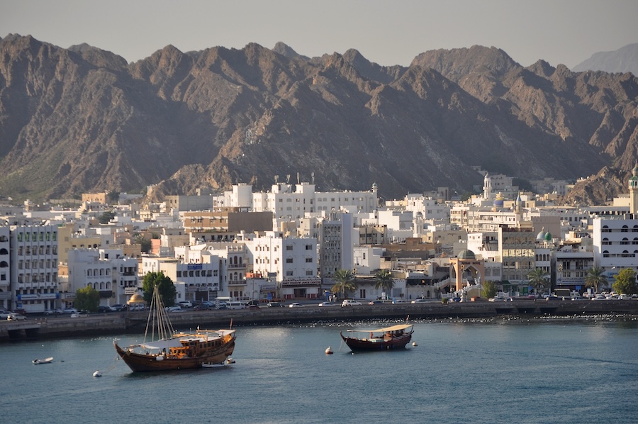 Muscat Harbor with white and beige buildings along a curved coastline, two wooden boats floating in calm blue water and rugged brown mountains rising in the background under a clear sky.