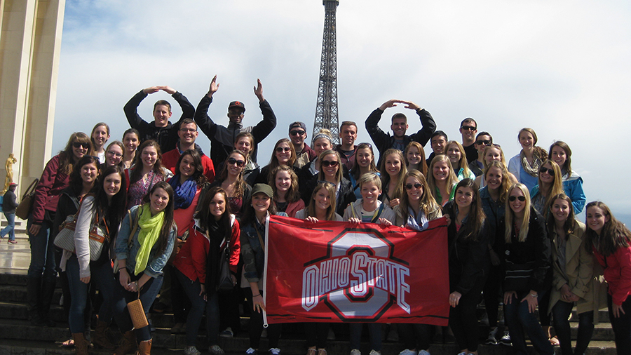 Large group of people standing together outdoors, holding an Ohio State flag, with the Eiffel Tower visible in the background.