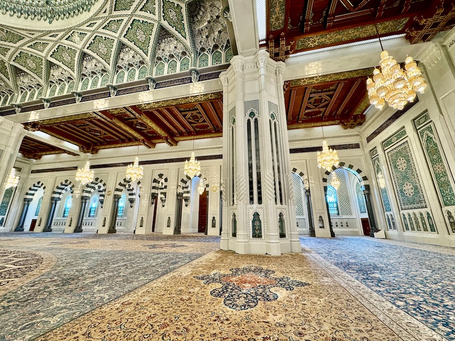 Oman Mosque interior with tall white columns, decorative arches, patterned ceilings, hanging chandeliers and a large patterned carpet covering the floor.
