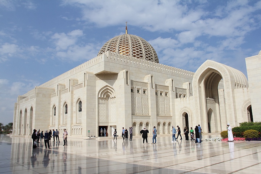 Oman Sultan Gaboos Grand Mosque with a patterned dome and arched entrances stands under a partly cloudy blue sky, with people walking across a wide reflective courtyard in front of the building.