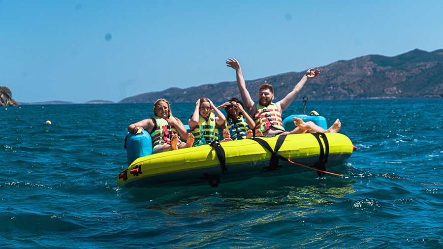 Group of people riding a yellow inflatable banana boat on blue ocean water, with distant mountains and a clear sky in the background.