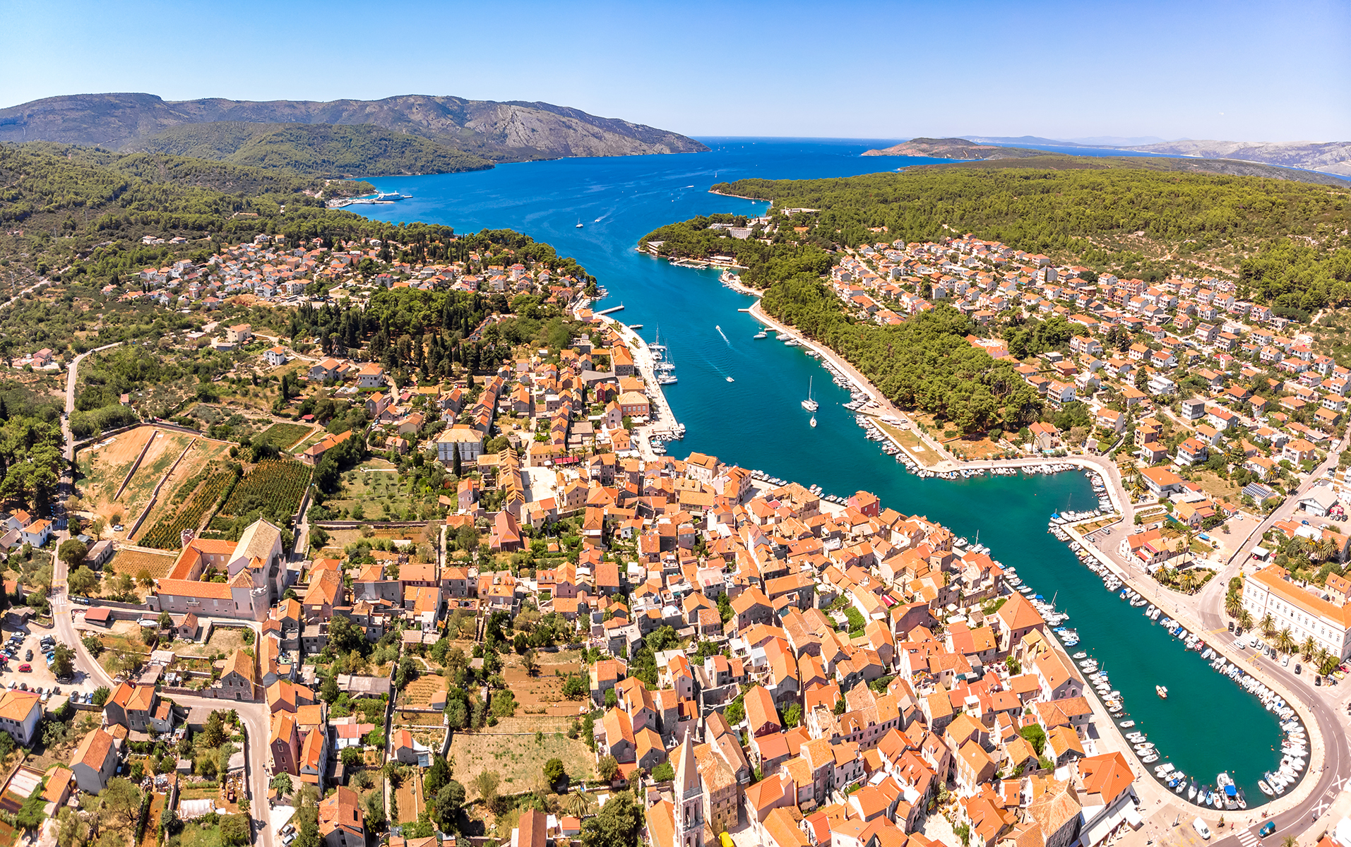 Aerial view of a coastal town with terracotta rooftops, a vibrant marina, lush green hills, and turquoise waters under a clear blue sky.
