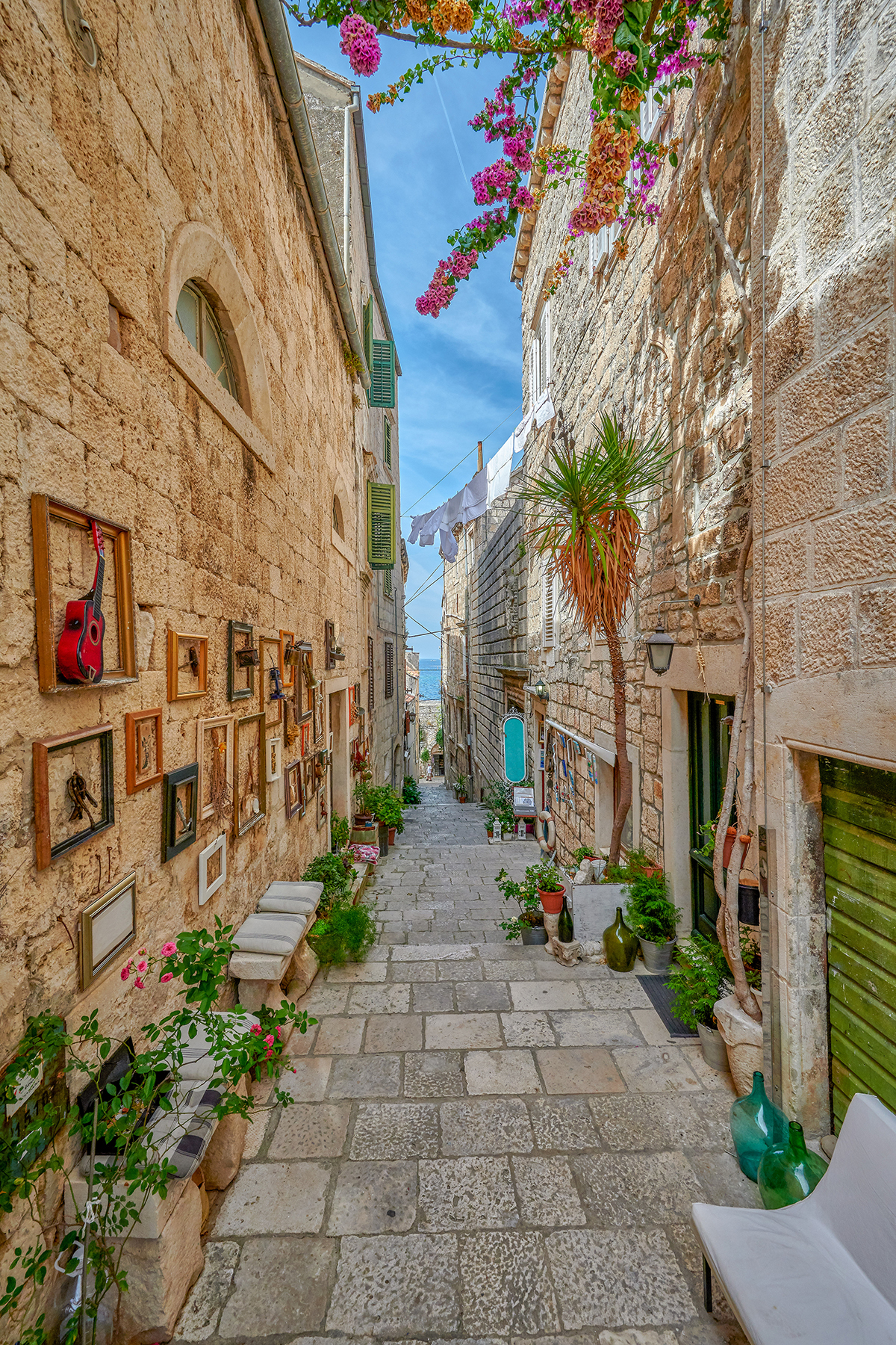 Charming narrow cobblestone alley adorned with framed artwork, potted plants, and colorful flowers under a sunny blue sky.