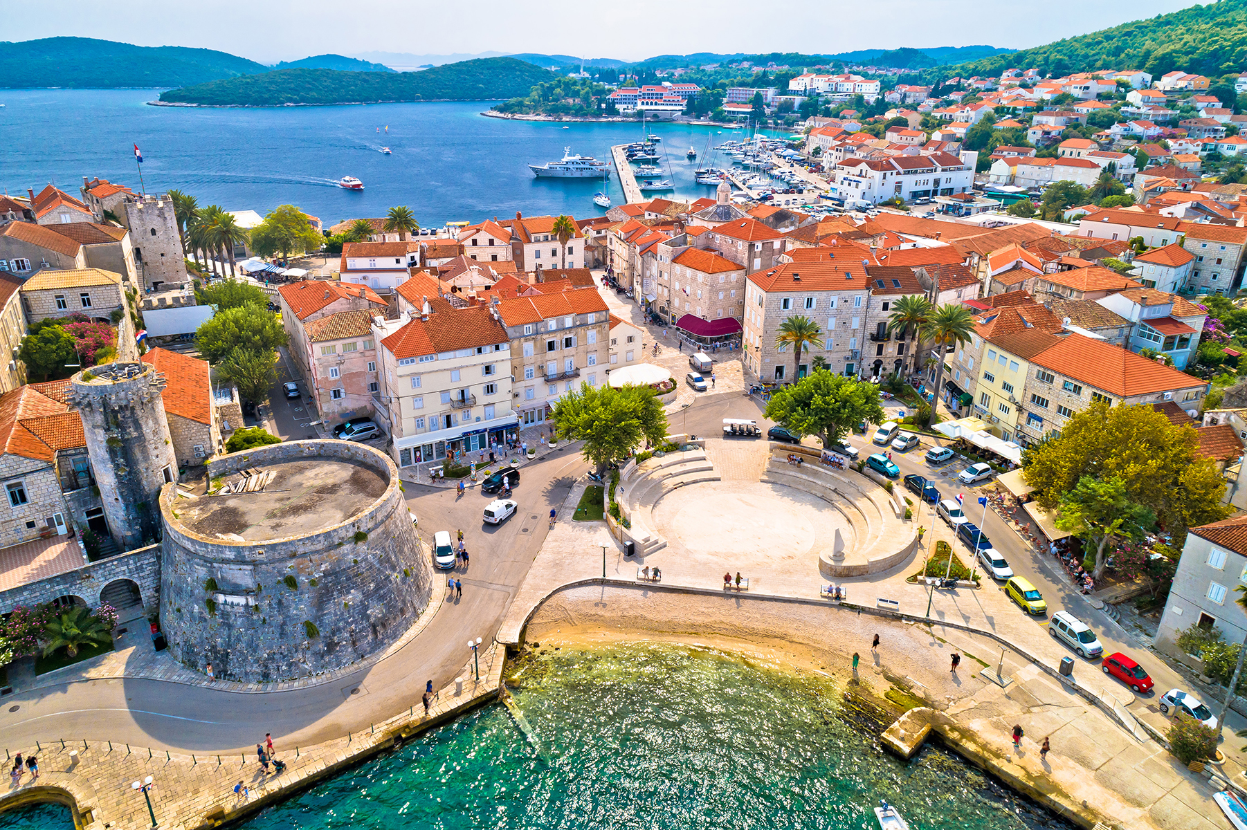 Aerial view of a coastal town with terracotta roofs, palm trees, a harbor, and a stone tower, surrounded by lush hills and turquoise water.