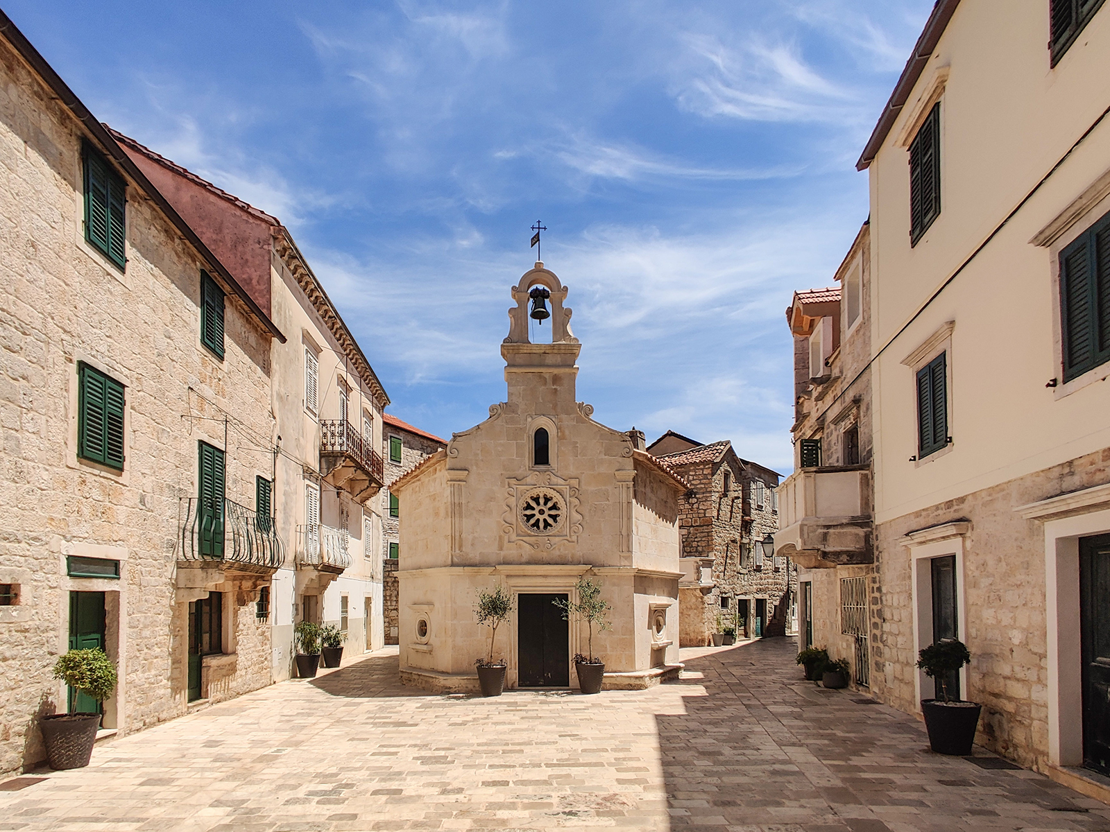 Charming stone buildings flanking a quaint square with a small church, under a blue sky with wispy clouds, inviting atmosphere.