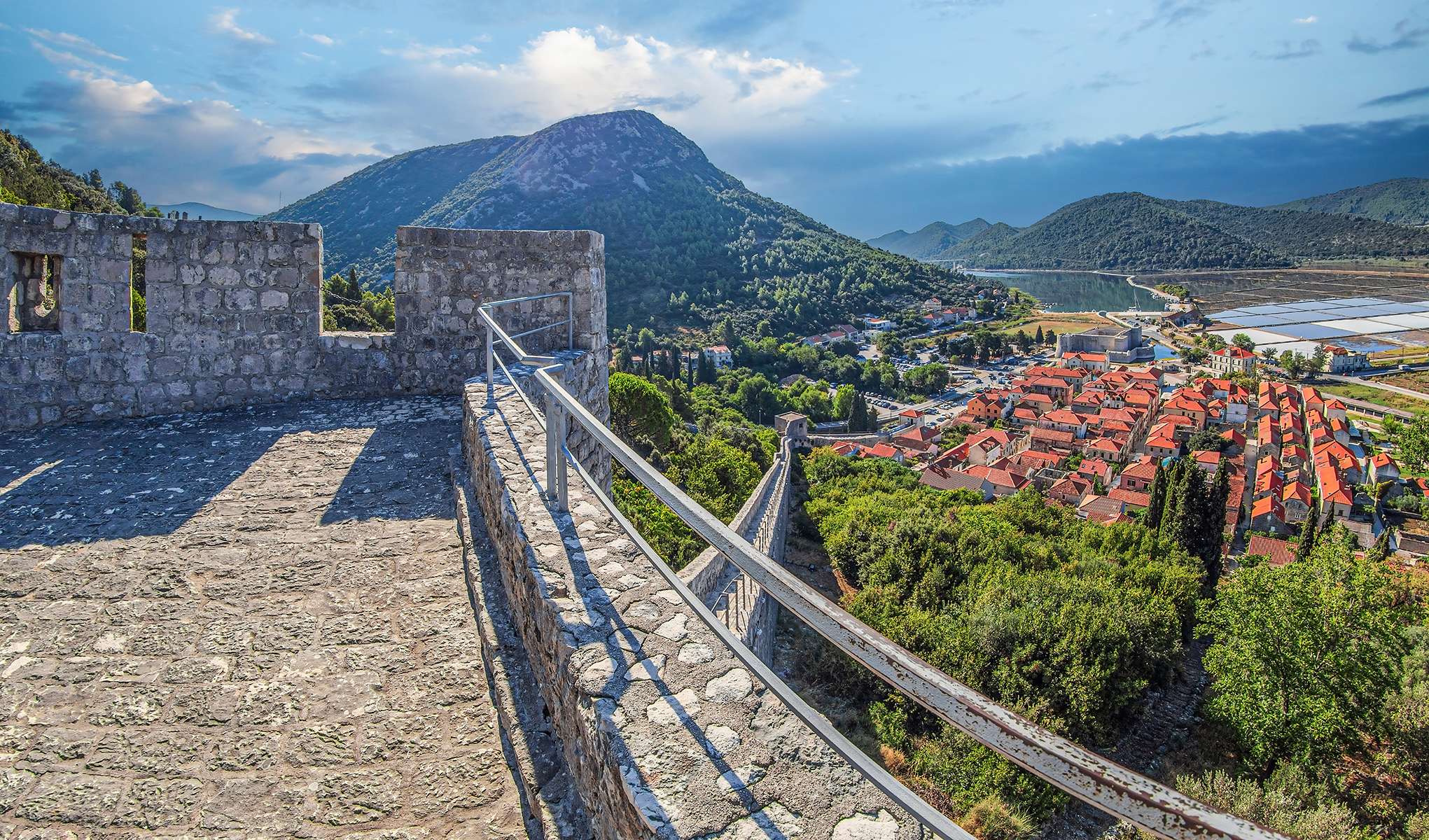 View from a stone castle overlook, featuring a mountainous landscape and a village with red-roofed houses below.
