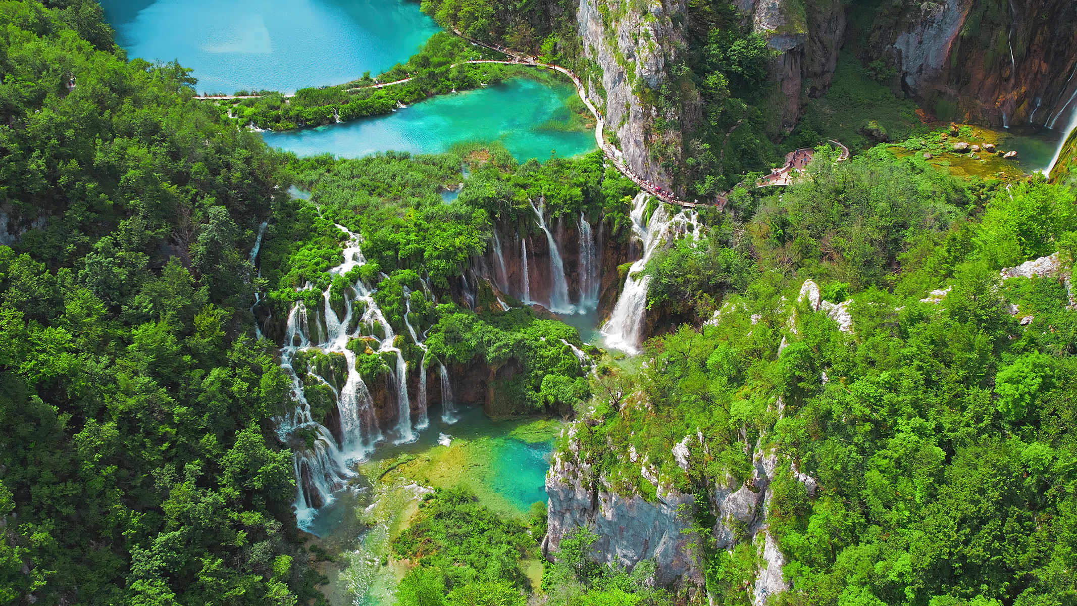 Aerial view of lush green waterfalls cascading into turquoise lakes, surrounded by dense foliage and walking paths in a natural landscape.