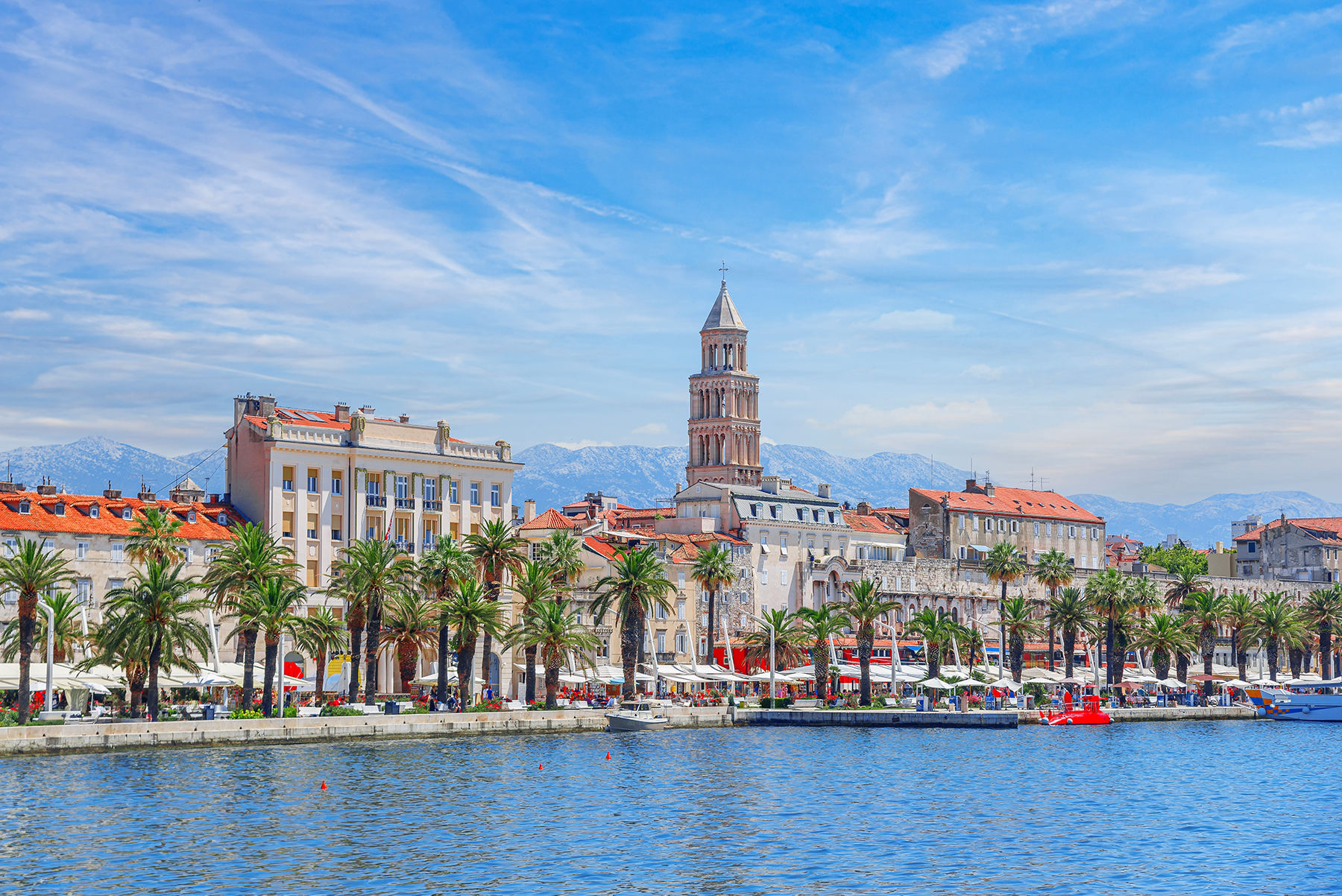 Coastal view of Split, Croatia, showcasing palm-lined promenade, historic buildings, and the iconic bell tower against a blue sky.