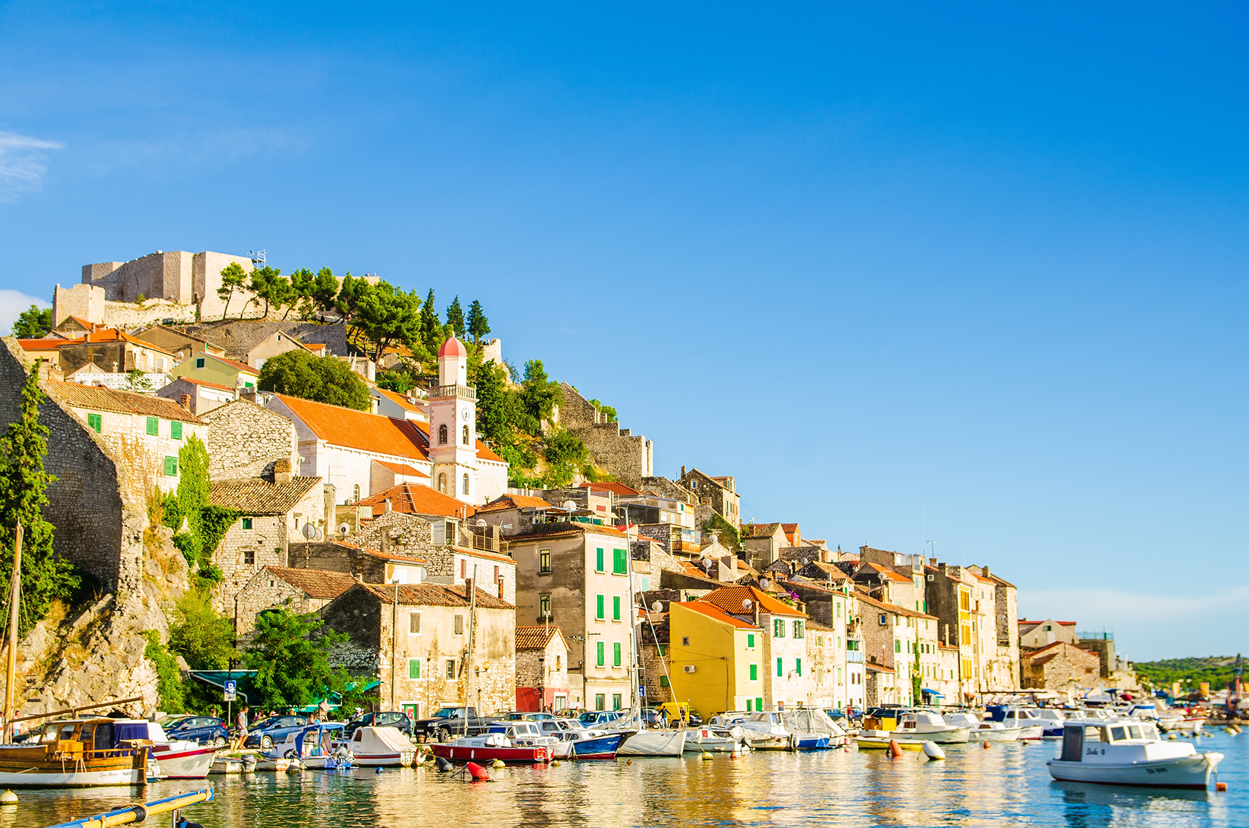 Charming coastal town with colorful buildings, boats in the harbor, and a historic castle overlooking a clear blue sky.
