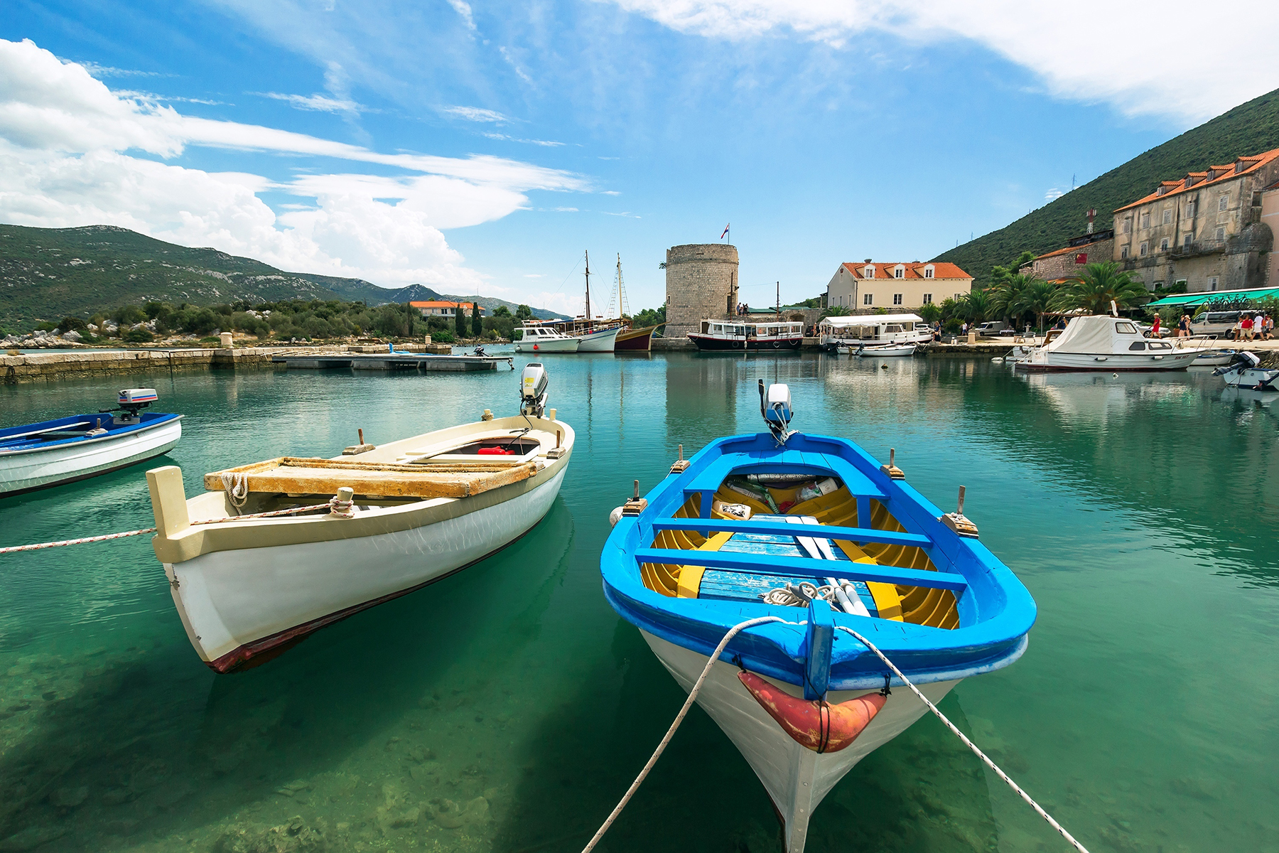 Colorful boats moored in a tranquil harbor, with green hills and a historic tower in the background under a bright blue sky.
