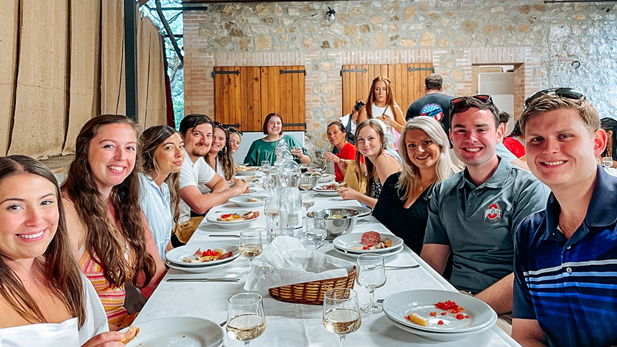 Long dining table set with plates and glasses, surrounded by a large group of people seated closely together inside a restaurant with stone walls and warm lighting.