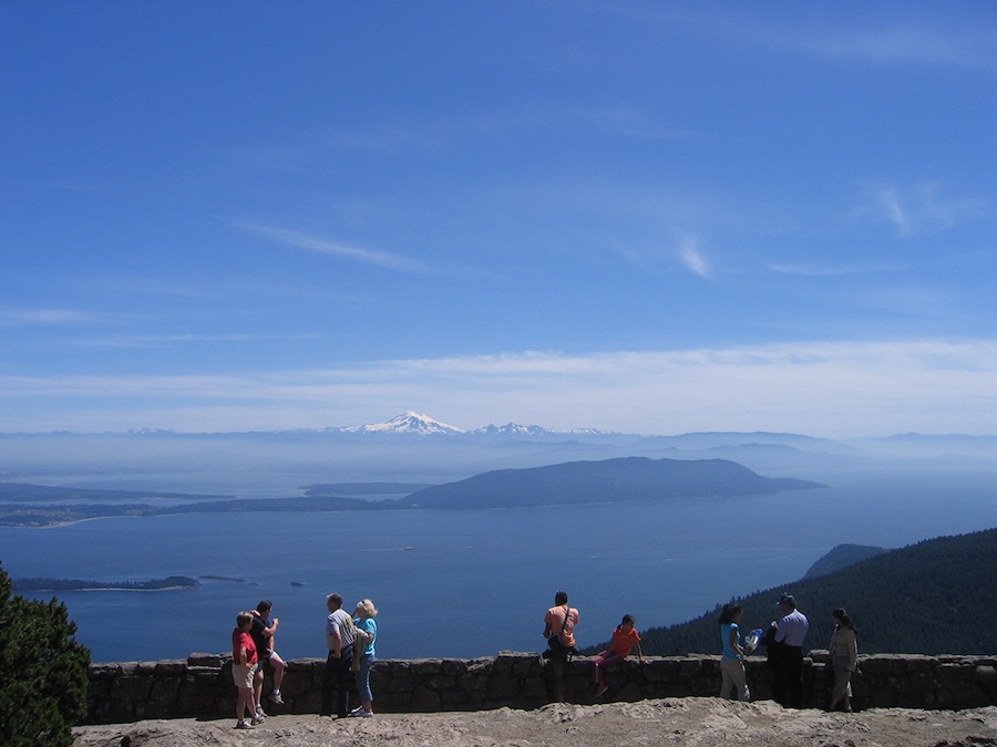 People standing at a stone overlook, looking out over blue water, forested islands, and a distant snow-capped mountain under a clear sky.