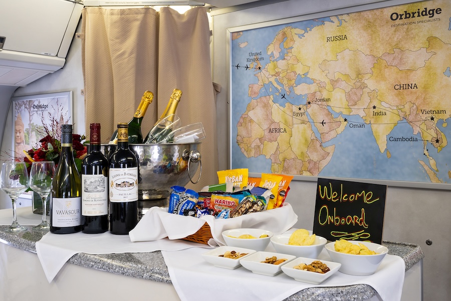 A small onboard buffet displays wine bottles, champagne, snacks, and bowls of food on a counter, with a world map on the wall and a sign that reads “Welcome Onboard.”