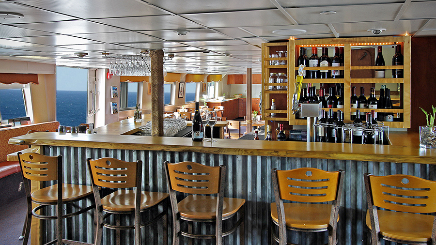A bar interior with wooden stools at a long counter, shelves of bottles behind the bar, and large windows letting in daylight.
