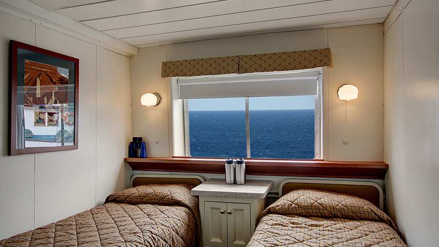 A small ship cabin with two single beds, a window showing ocean water, a built-in desk between the beds, and wall lights on either side.
