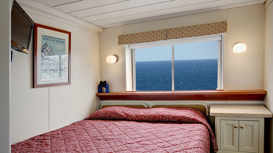 A small ship cabin with one double bed, a window showing ocean water, a built-in desk, and wall lights on either side of the bed.