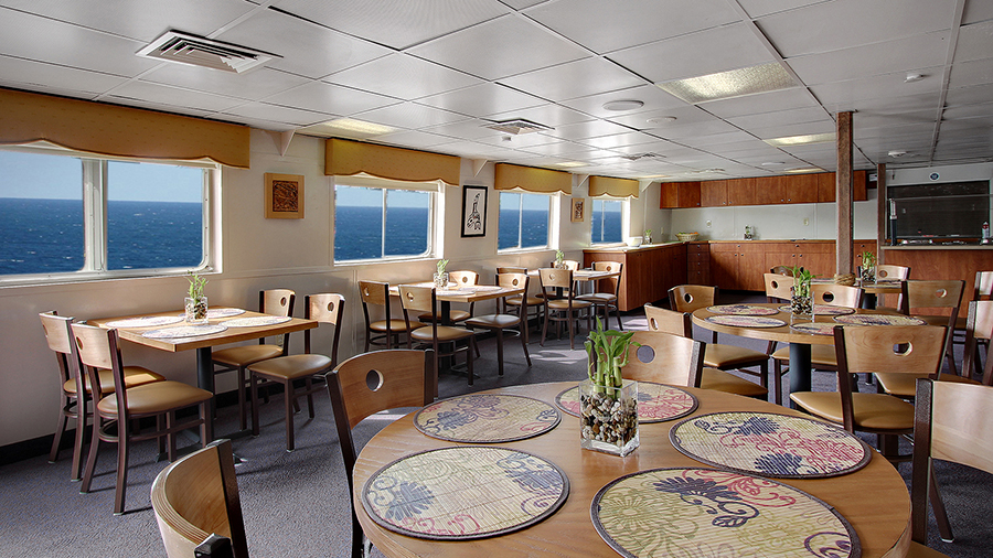 A bright dining room with wooden tables and chairs, round placemats on the tables, large windows along the walls, and a serving area in the background.