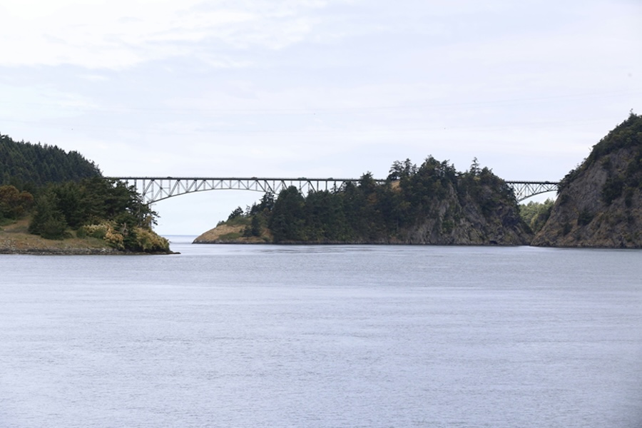 A wide view of calm water between two forested rocky islands, with a steel bridge spanning the gap in the distance.