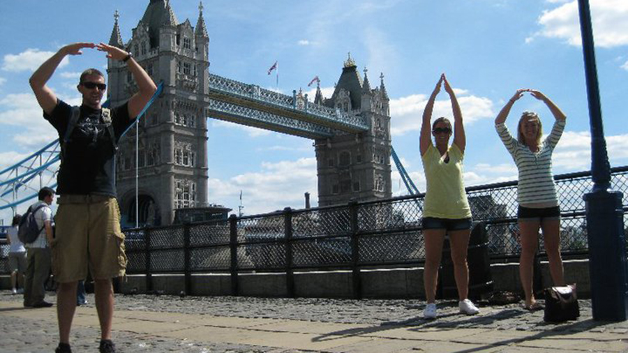 Three people standing on a riverside walkway with arms raised in circle shapes, with Tower Bridge and the River Thames visible behind them.