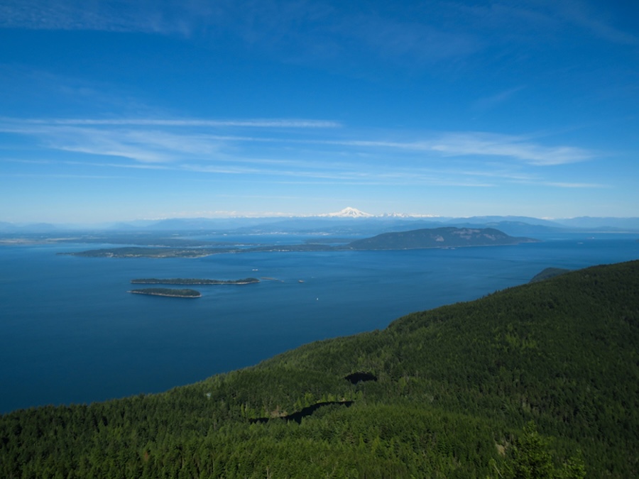 An aerial view of blue ocean and small islands, with forested hills in the foreground and a distant snow-capped mountain under a clear sky.