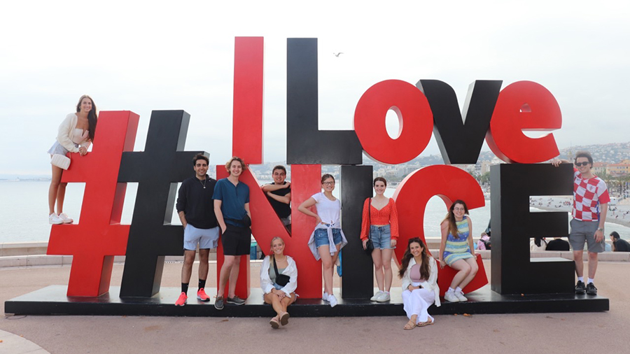 Group of people posing in front of a large outdoor sign reading “#ILoveNICE,” with red and black letters on a paved plaza.