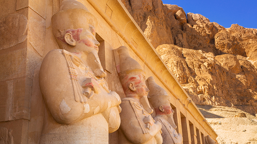 Ancient Egyptian statues with headdresses line a sunlit stone wall, backed by rugged cliffs and a clear blue sky, conveying historical grandeur.