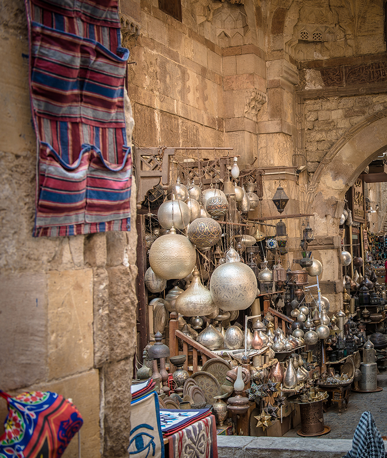 A vibrant Middle Eastern market stall displaying ornate silver lamps and decorations. The scene is framed by stone archways and colorful, patterned fabrics.