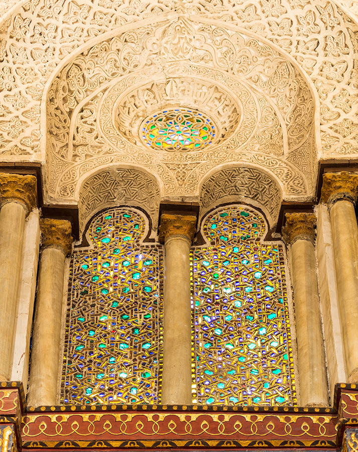 Ornate architectural detail with three arched stained glass windows framed by stone columns. Decorative patterns and warm light create a serene atmosphere.