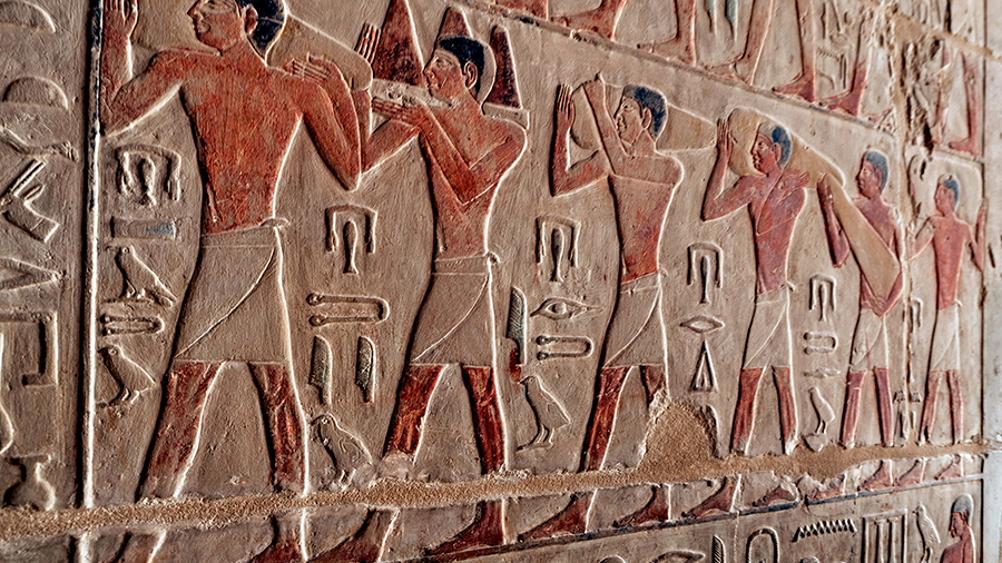 Ancient Egyptian wall relief depicts six men in profile, wearing kilts, carrying objects. Hieroglyphs surround them. Earthy tones convey a historical scene.